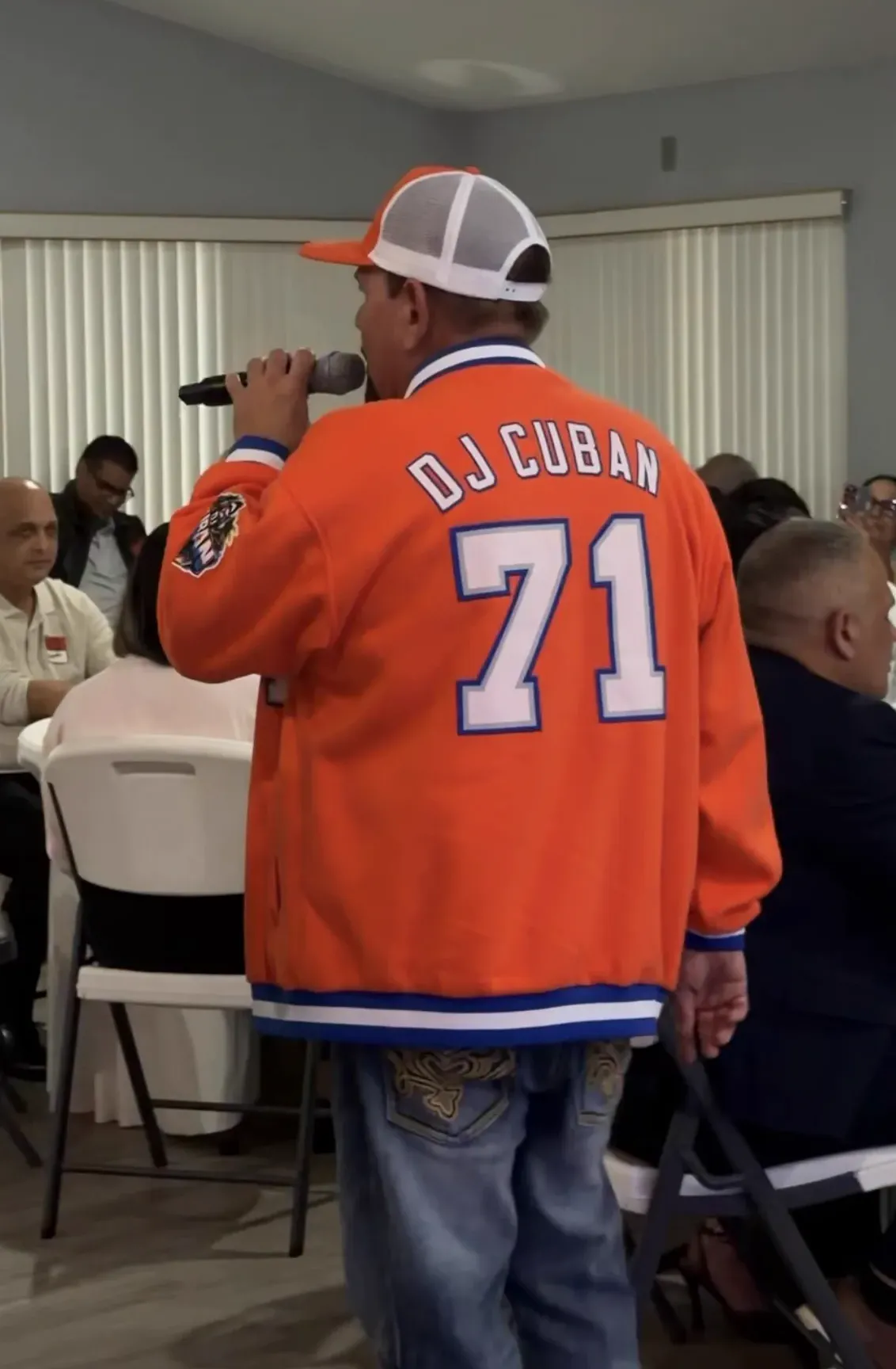 DJ Cuban singing to seated guests, in his orange DJ Cuban 71 jersey