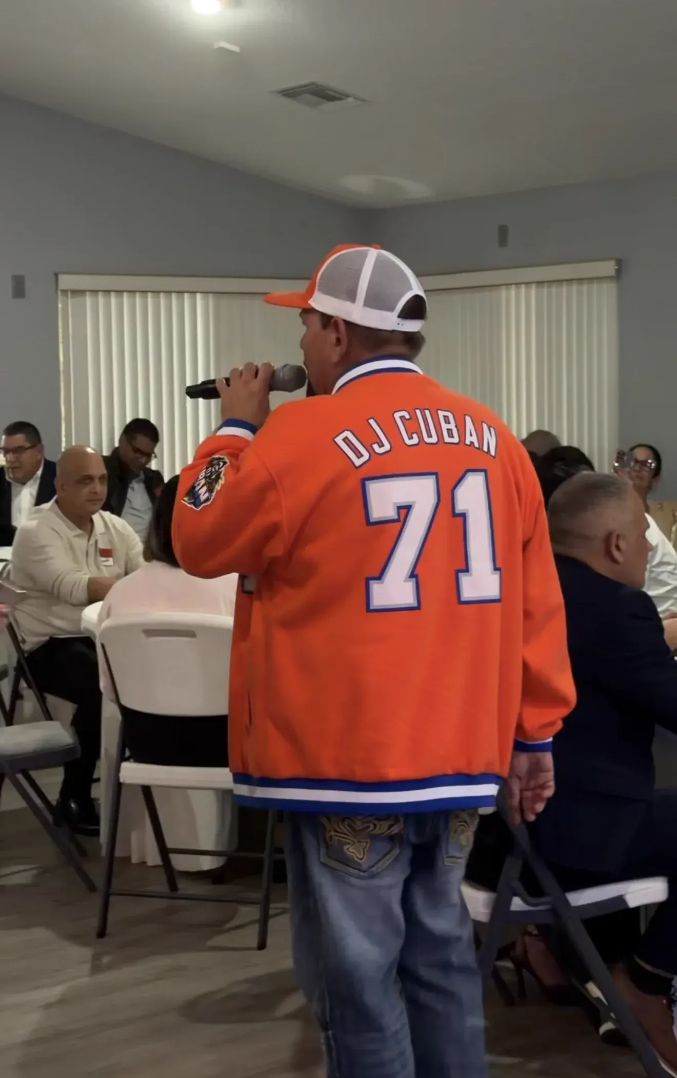 DJ Cuban singing to seated guests, in his orange DJ Cuban 71 jersey